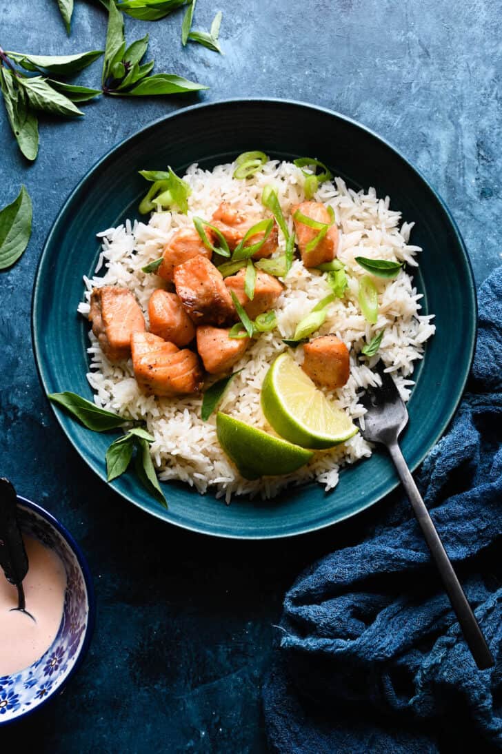 A blue bowl filled with white rice, air fryer salmon bites, sliced green onions and lime wedges, with a fork sticking into the rice.