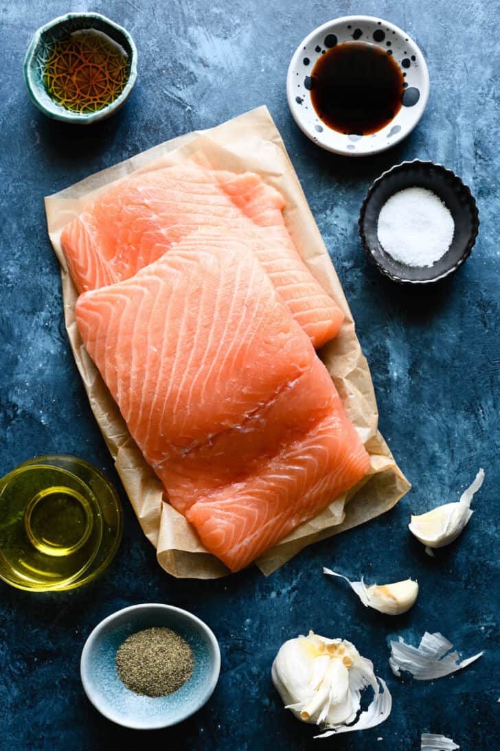 Ingredients laid out on a blue surface, including fillets of pink fish, soy sauce, oil and salt and pepper.