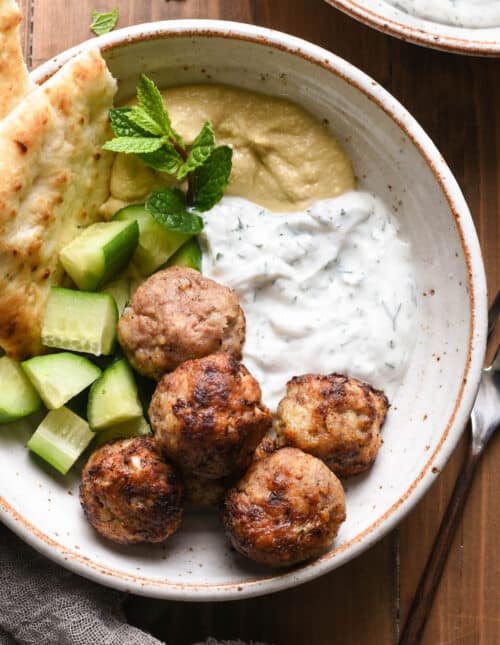 Overhead photo of rustic bowl filled with air fryer turkey meatballs, chopped cucumber, hummus, yogurt sauce, naan and a mint sprig garnish.