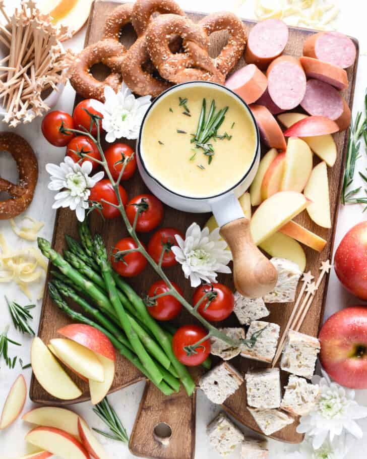 A unique charcuterie board featuring cheese fondue, smoked sausage, veggies, bread, pretzels and apples.
