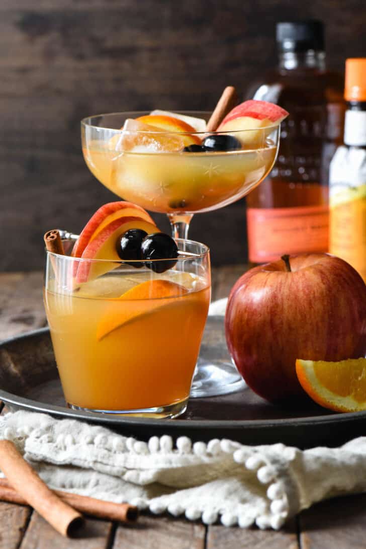 Bourbon apple cider drinks in two different glasses (1 tall, 1 short), on a tray with a whole apple.
