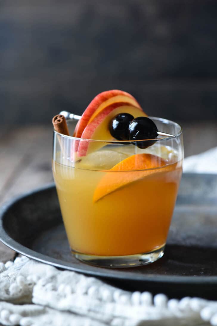 A fall cocktail in a small glass, garnished with an orange slice, cinnamon stick, and metal skewer with apple slices and cocktail cherries.