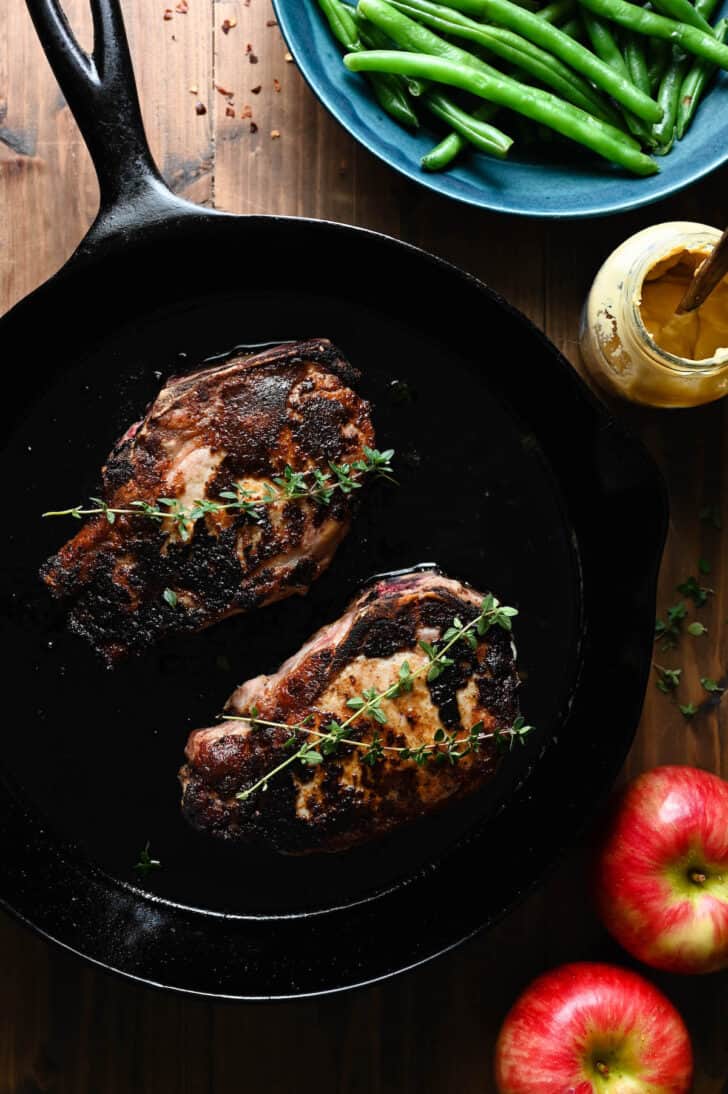 Two cooked pork chops in a cast iron skillet garnished with thyme.