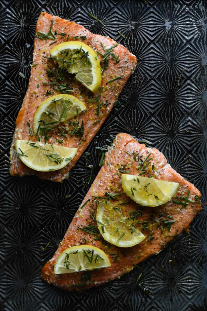 Baked orange fish fillets covered in herbs and lemon slices on a textured metal baking pan.