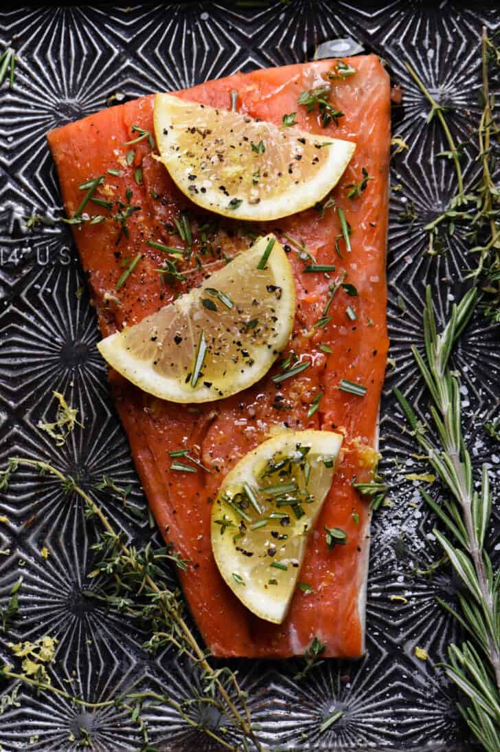 A fillet of sockeye salmon topped with lemon slices and herbs on a textured baking pan.