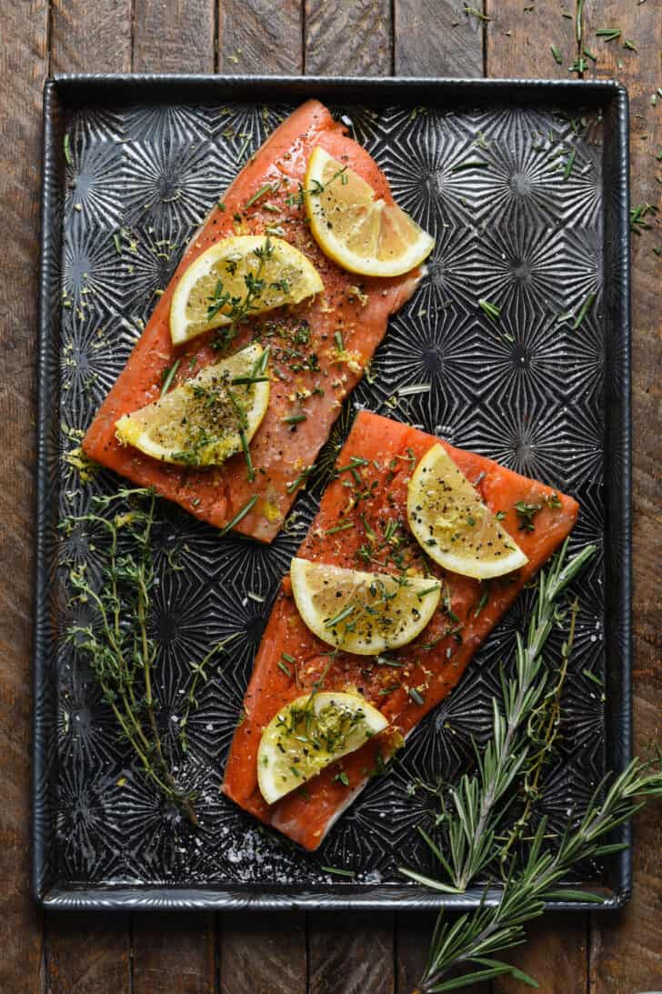 Sockeye salmon fillets topped with lemon slices and herbs on a textured baking pan.