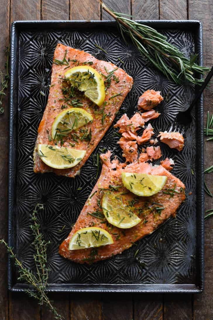 Baked sockeye salmon covered in herbs and lemon slices, being flaked with a fork on a textured baking pan.