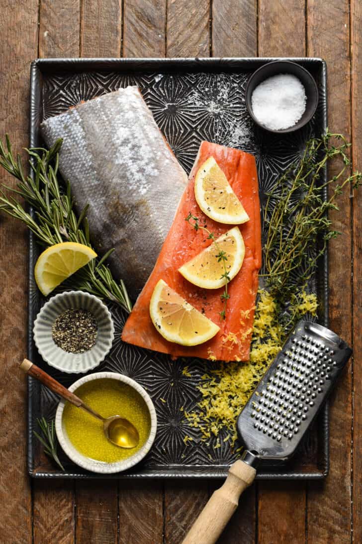 A textured baking pan filled with the ingredients needed for baked sockeye salmon, including fish, lemon, herbs, salt and pepper.