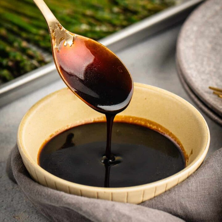 A gold spoon dripping balsamic glaze into a small beige bowl, with a tray of roasted asparagus in the background.