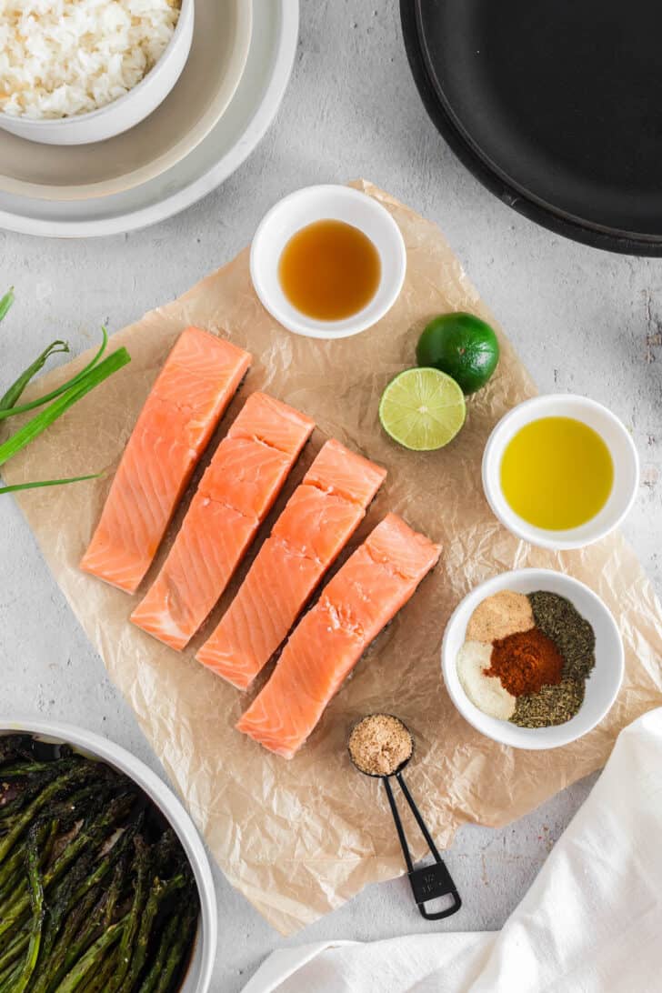 Ingredients laid out on a light surface including fish, spices, brown sugar and a lime.