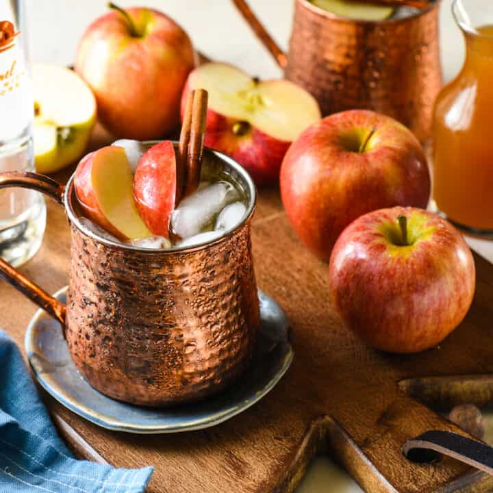 Wooden cutting board topped with apples and copper mugs filled with apple cider mule cocktails.