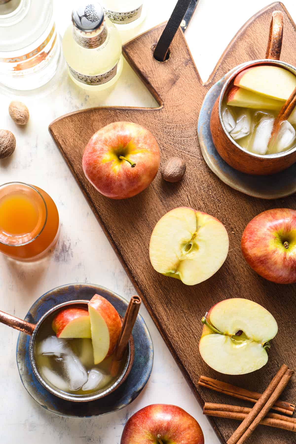 Overhead shot of a wooden cutting board with cut and whole apples, nutmeg pods, bottles, cinnamon sticks, and copper mugs filled with caramel apple cider mule cocktails.
