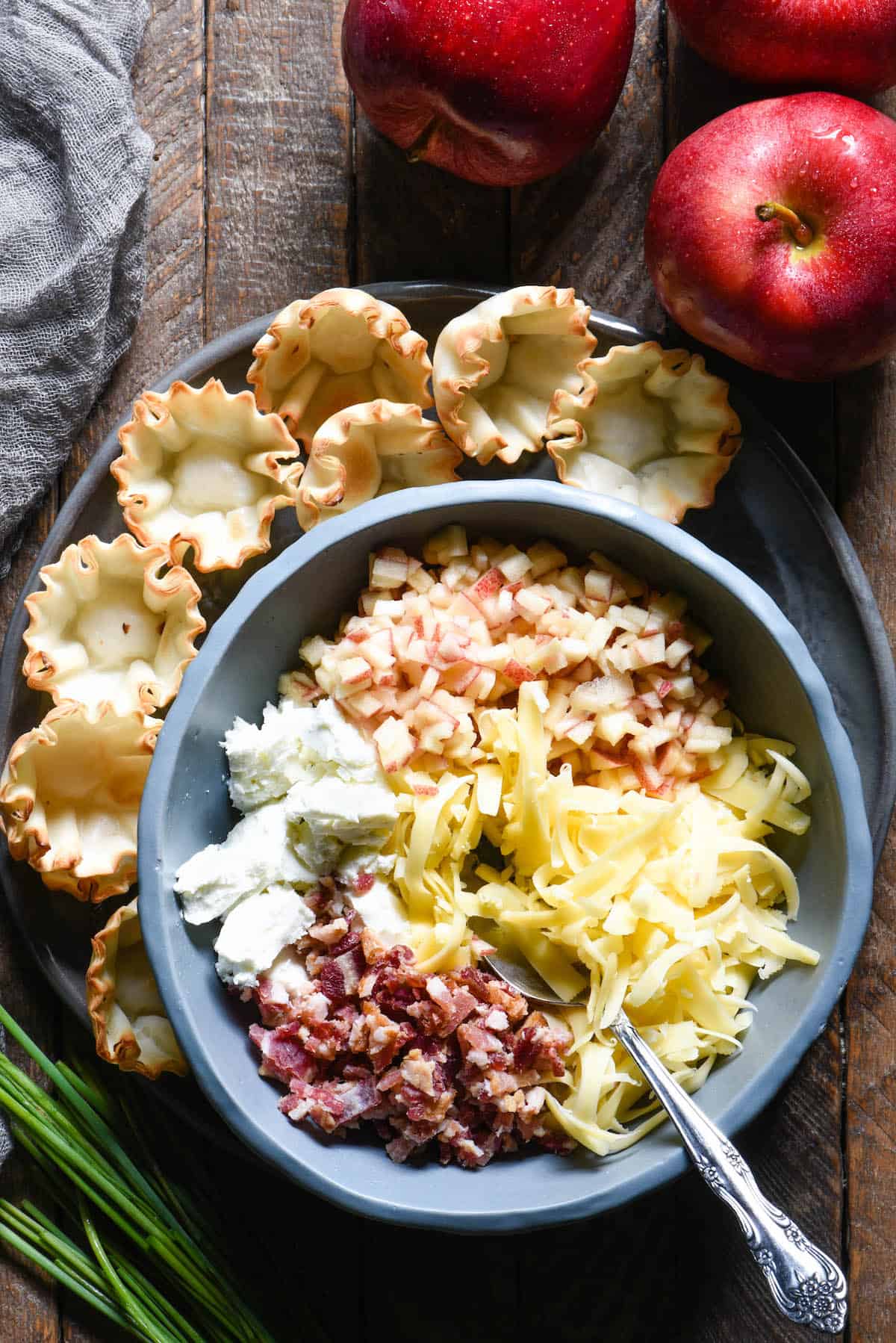 Bowl filled with bacon, goat cheese, light yellow shredded cheese and chopped apples, with empty phyllo cups nearby.