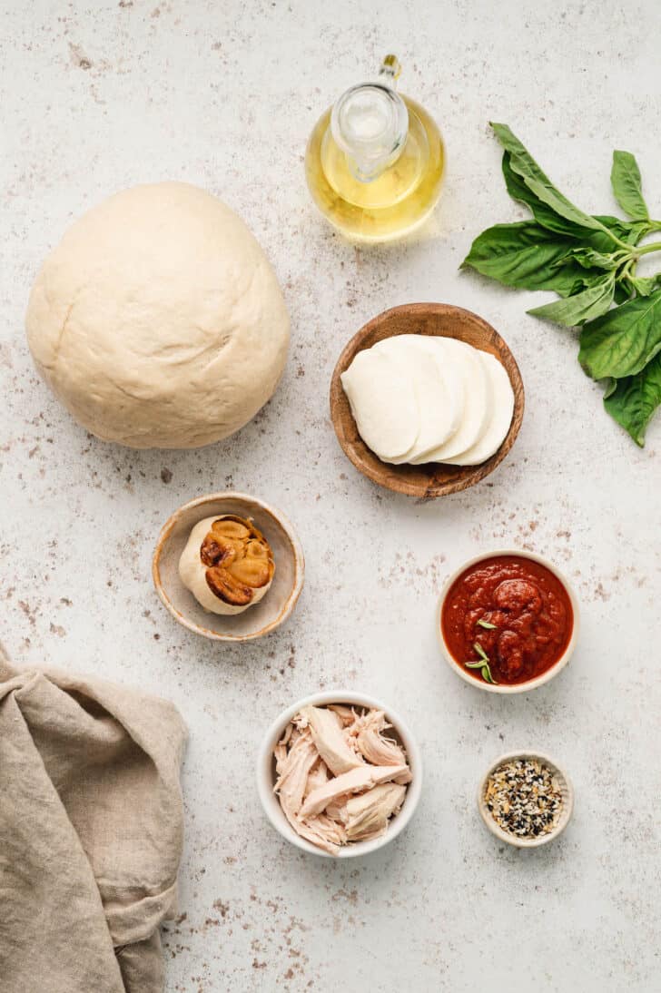 Ingredients needed for a chicken pizza recipe laid out on a light surface, including pizza dough, cheese, roasted garlic, pizza sauce, basil, oil, chicken and spices.