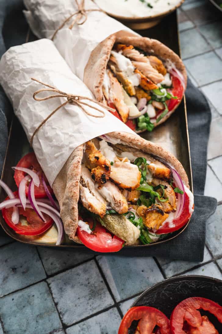 Two grilled chicken shawarma wraps, wrapped in white paper, on a gold platter on a tiled surface.