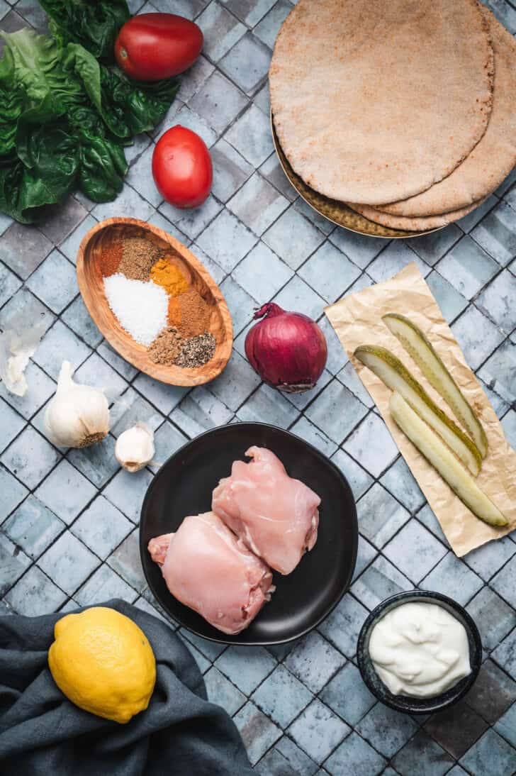 Ingredients laid out on a tile surface, including raw poultry, spices, onion, garlic, yogurt, pickles, pita bread, lettuce and tomato.