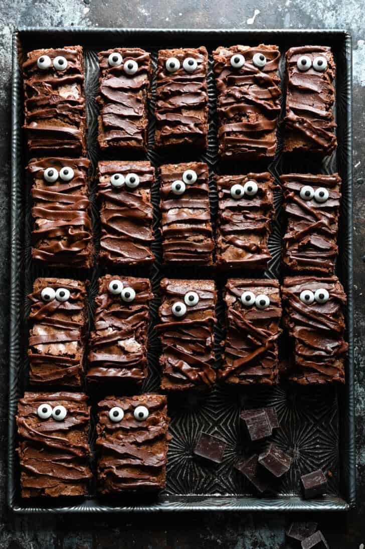 Brownies with chocolate ganache stripes and candy eyeballs on a textured baking pan.