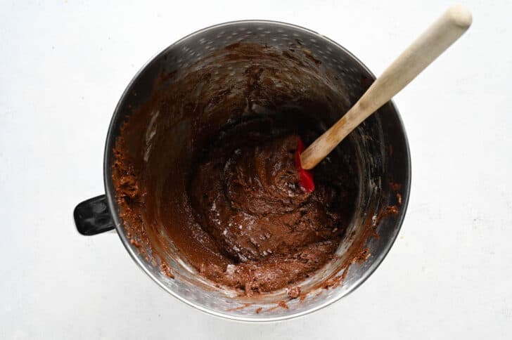 A red rubber spatula stirring a chocolate batter mixture in the bowl of a stand mixer.