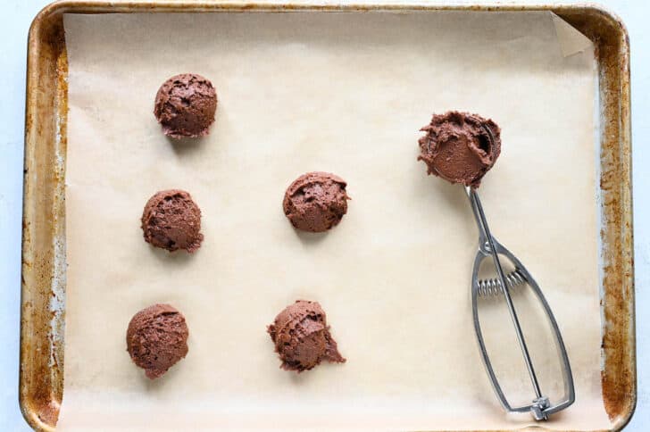 A cookie scoop scooping portions of chocolate cookie dough onto a baking pan lined with parchment paper.
