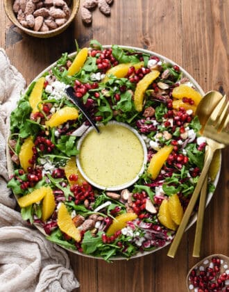 A Christmas Salad made with greens, oranges, cheese, pomegranate seeds and nuts, arranged like a wreath with a bowl of dressing in the middle.