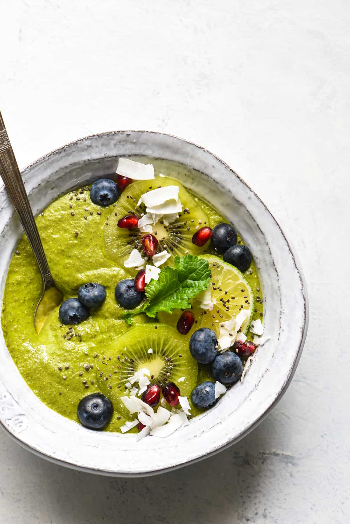 A green smoothie bowl topped with blueberries, pomegranate seeds, coconut, kiwi and a green leaf.