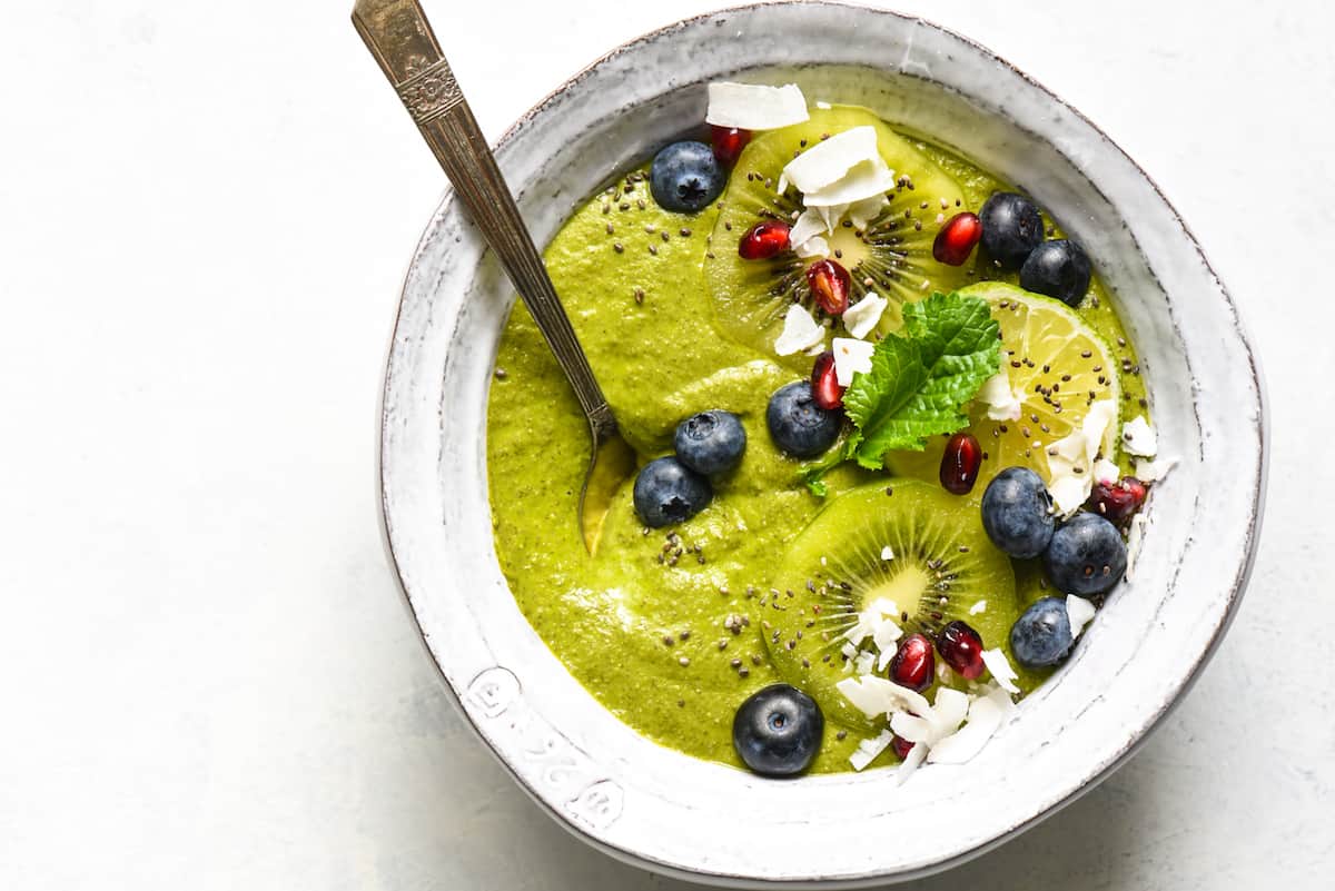 A green smoothie bowl topped with blueberries, pomegranate seeds, coconut, kiwi and a green leaf.