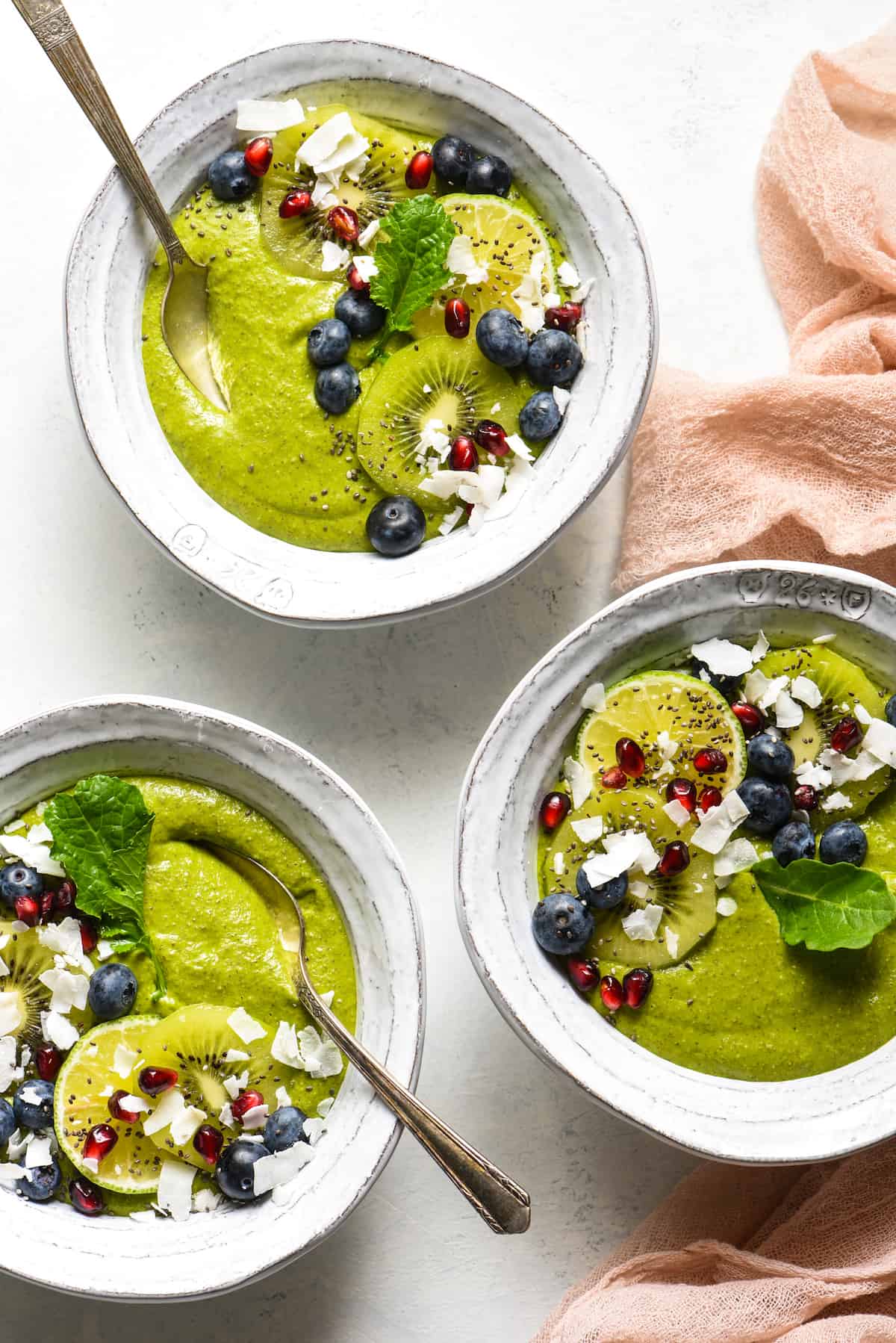 Three fruit smoothie bowls topped with blueberries, pomegranate seeds, coconut, kiwi and a green leaf.