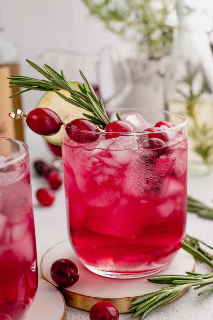 A cranberry rosemary cocktail garnished with a rosemary sprig, fresh cranberries and a wheel of lime.