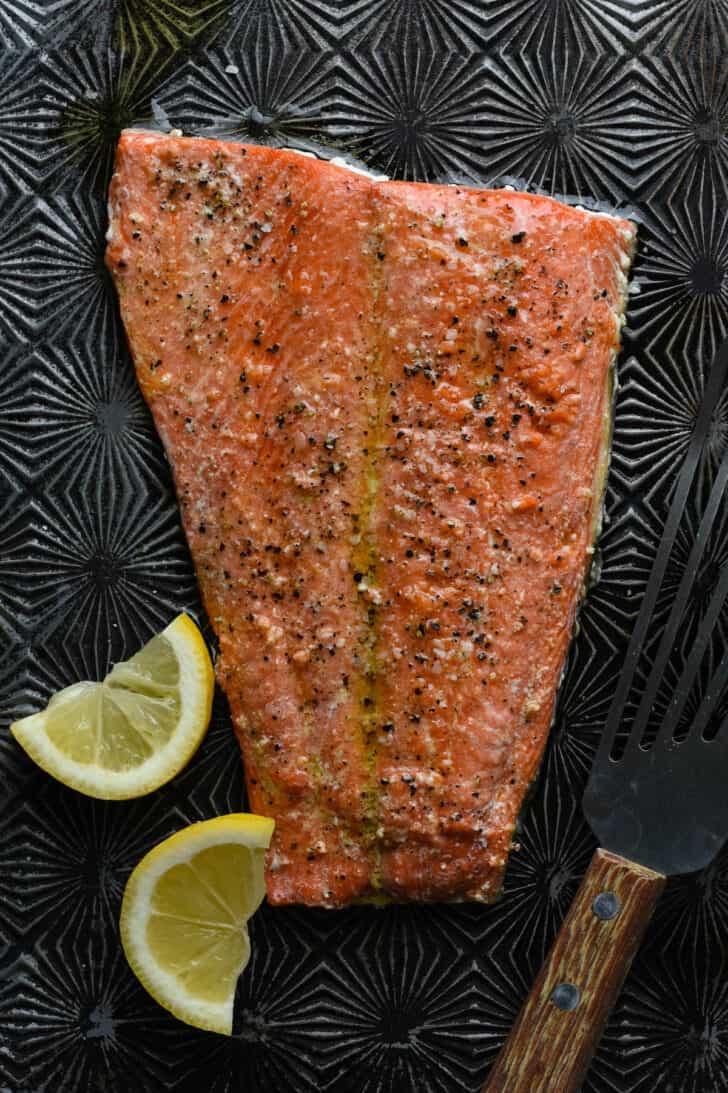 A large fillet of baked sockeye salmon on a textured baking pan with lemon wedges on the side.