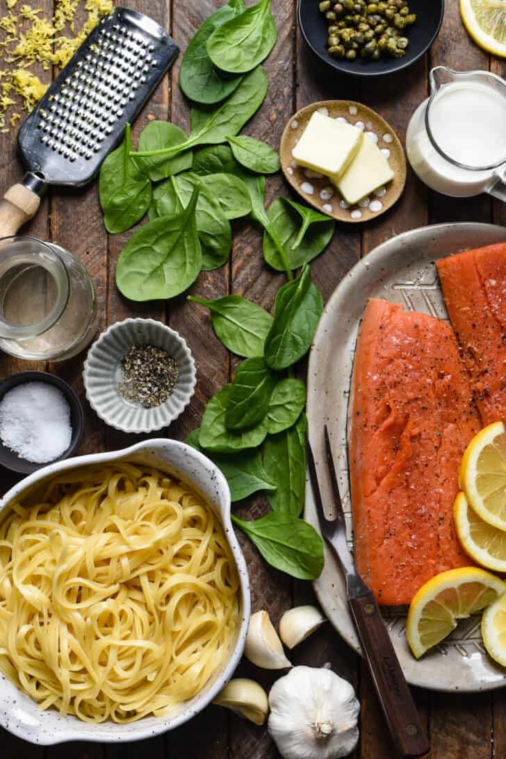 Ingredients laid out on a wooden surface, including linguine, fish with bright orange flesh, garlic, spinach, butter, capers, spices, cream and lemon zest.