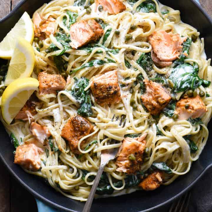 A large blue cast iron skillet filled with creamy salmon pasta with spinach, and lemon wedges as garnish.