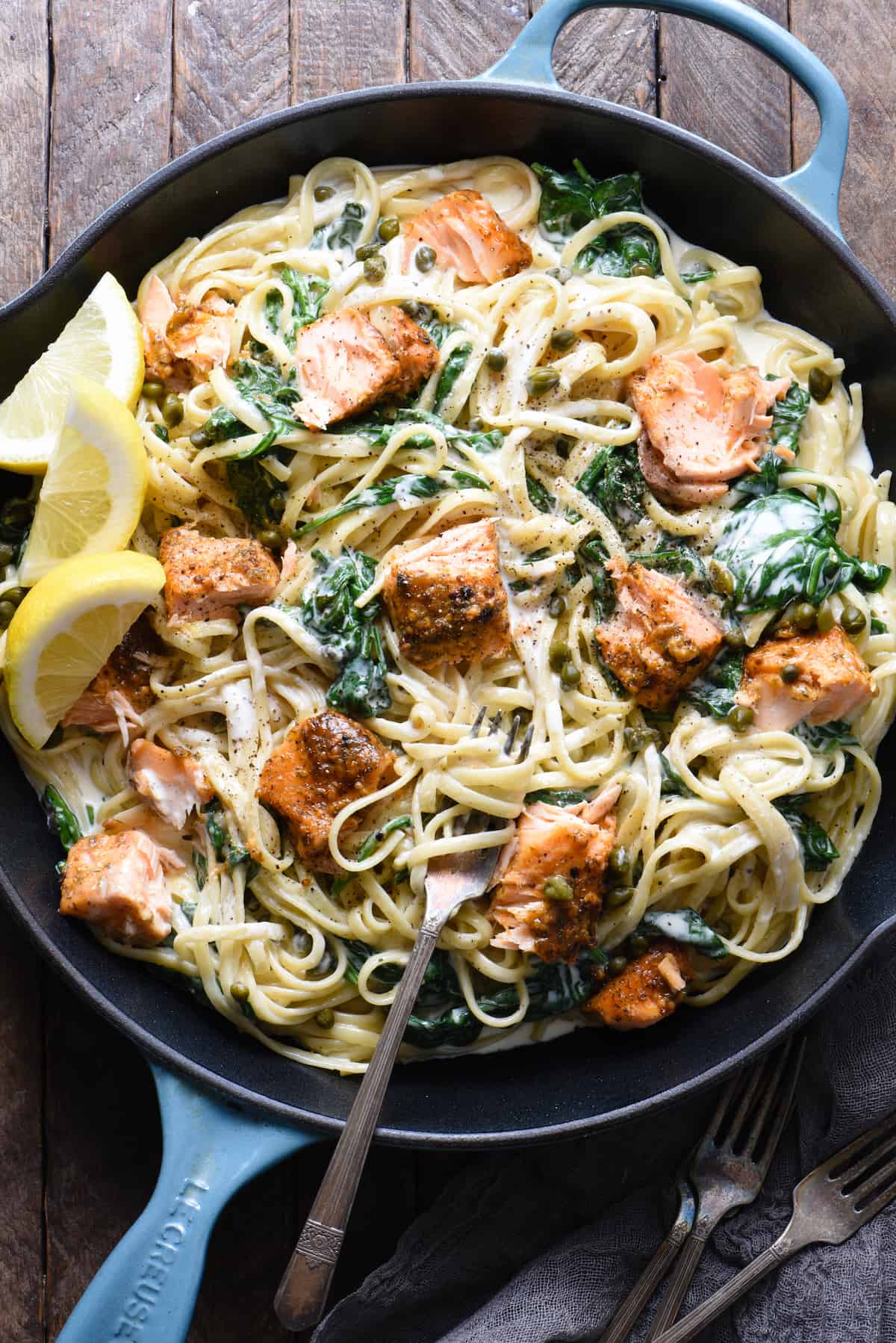 A large blue cast iron skillet filled with creamy salmon pasta with spinach, and lemon wedges as garnish.