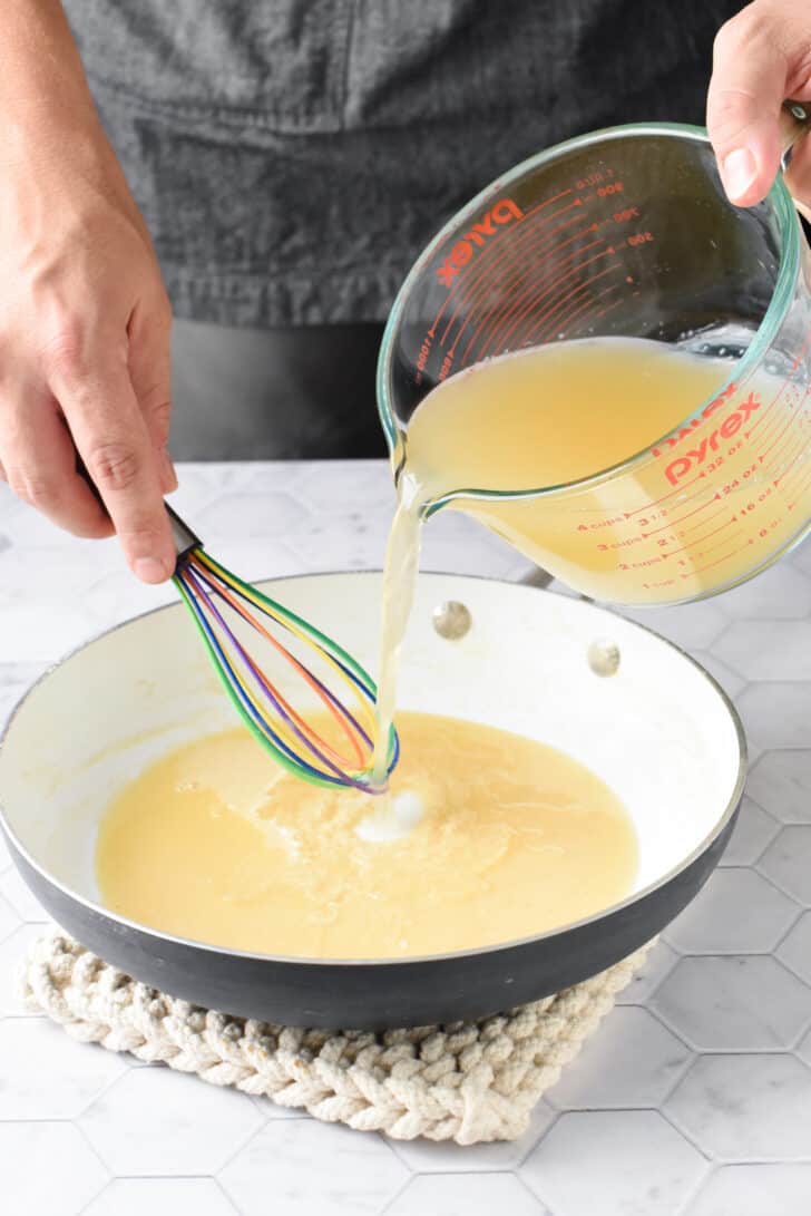 A man's hands adding broth from a large glass measuring cup to a white skillet with roux in it, while whisking with a colorful whisk.