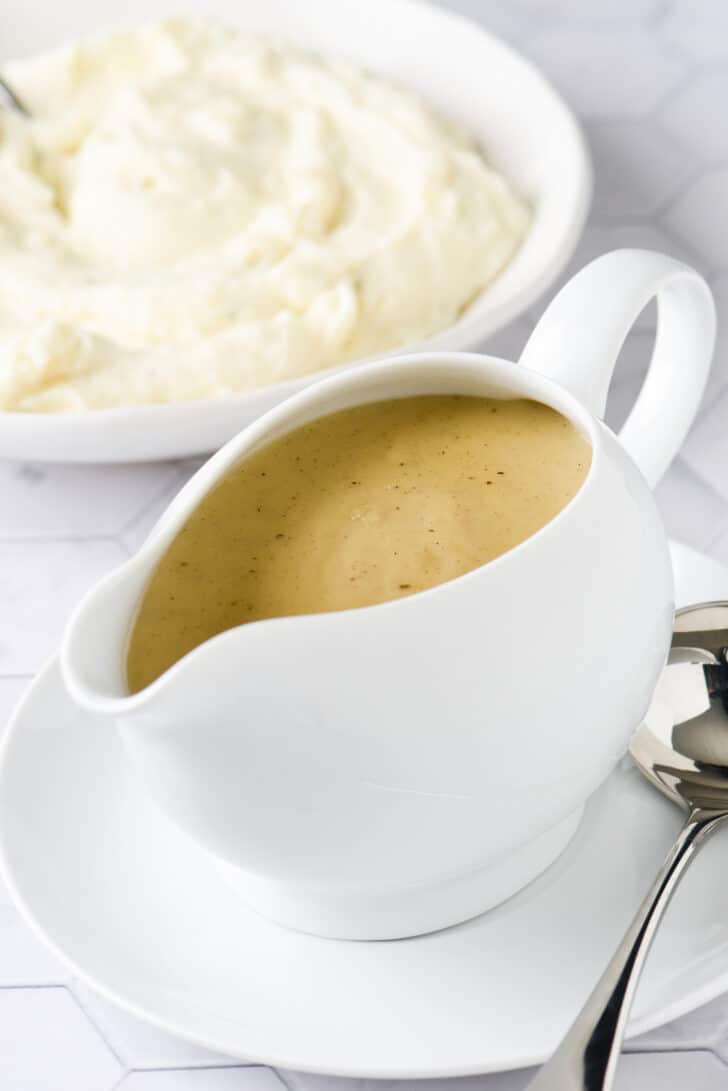 White ceramic boat filled with homemade gravy, with bowl of mashed potatoes in background.