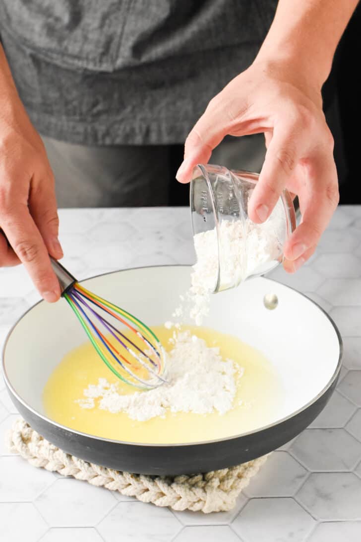 A man's hands adding a small ramekin of flour to melted butter in a white skillet, while whisking with a multicolored whisk.