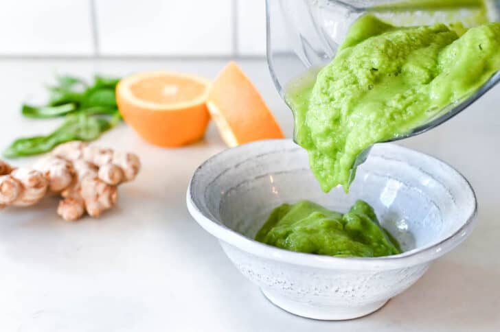 A green smoothie bowl mixture being poured from a blender pitcher into a gray bowl.