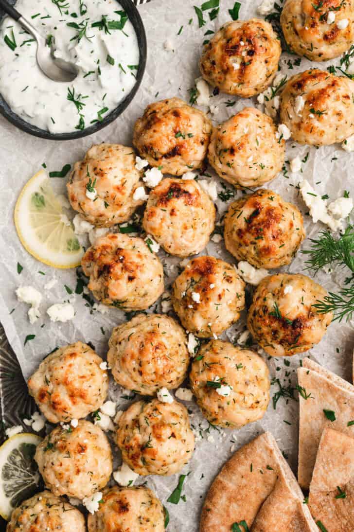 Greek chicken meatballs on white parchment paper, with crumbled feta, fresh herbs, lemon slices and yogurt sauce.