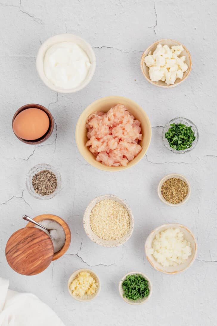 Ingredients in small bowls on a white surface, including ground poultry, feta cheese, breadcrumbs, an egg, onions, and herbs and spices.