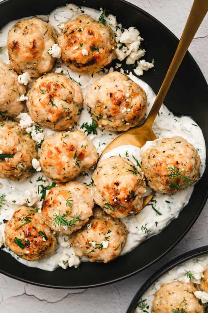 A black bowl filled with herby yogurt sauce and Greek chicken meatballs, with a spoon digging in.