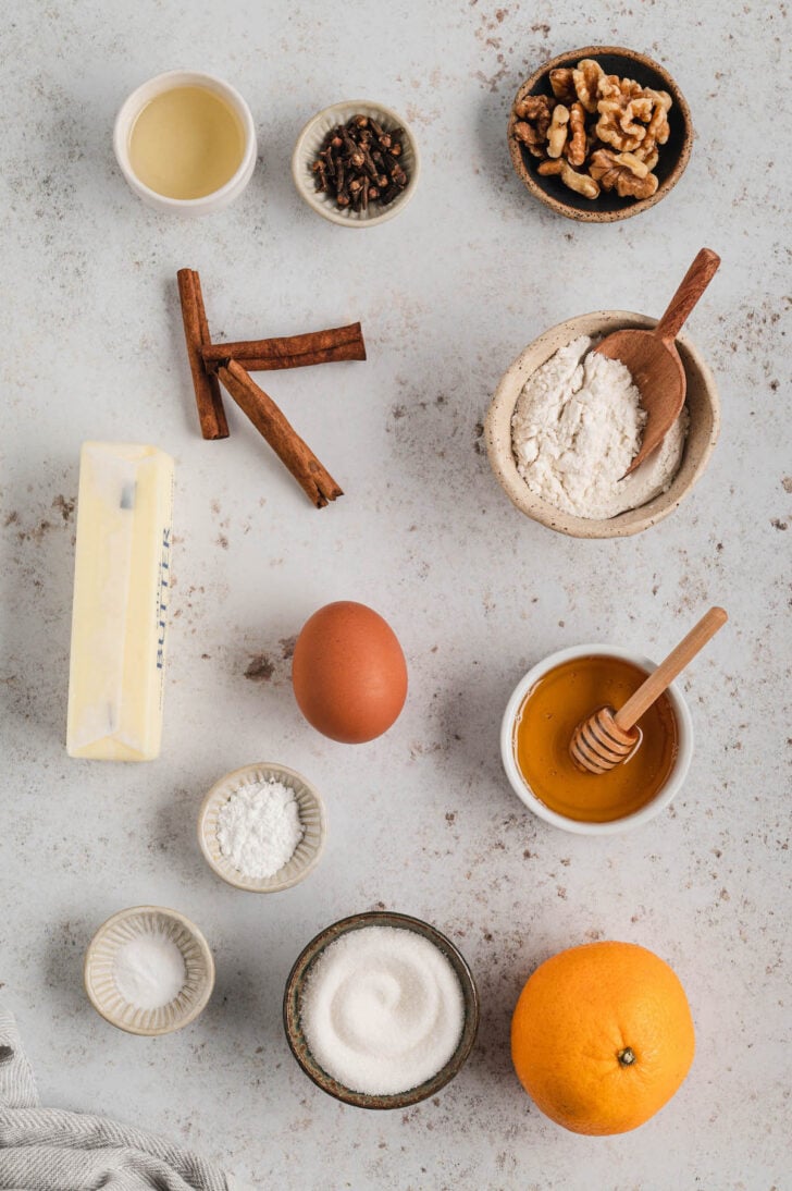 Ingredients on a light surface, including flour, sugar, butter, spices, honey, walnuts and an orange.