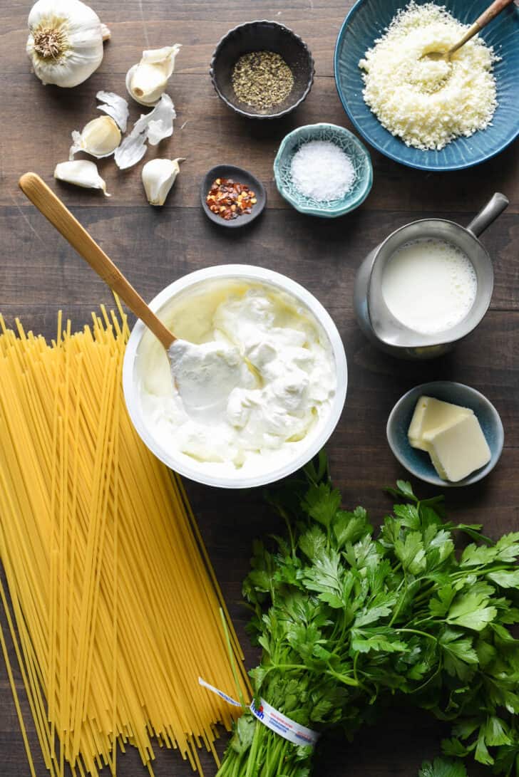 Ingredients laid out on a wooden table, including spaghetti, yogurt, butter, milk, spices, garlic, parsley and Parmesan cheese.