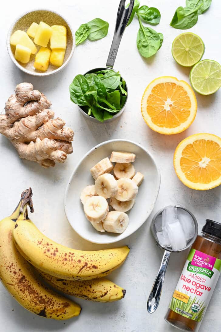 Ingredients arranged on a light surface, including bananas, fresh ginger, oranges, agave nectar, limes, pineapple and spinach.