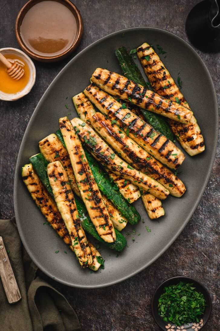 Grilled zucchini spears with a miso glaze on a dark gray oval platter with a serving fork.