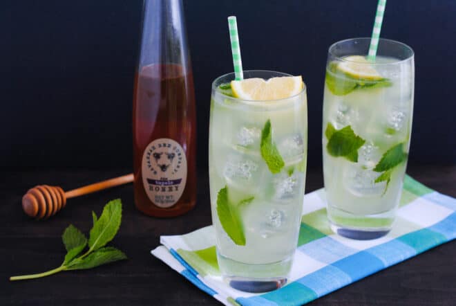 Two tall glasses of lemon mint drink with a bottle of honey and honey dripper in background.