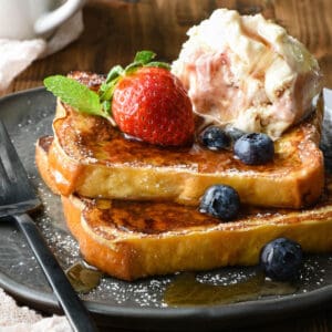 Two slices of ice cream French toast on a gray plate, topped blueberries, mint, a strawberry, a scoop of ice cream and maple syrup.