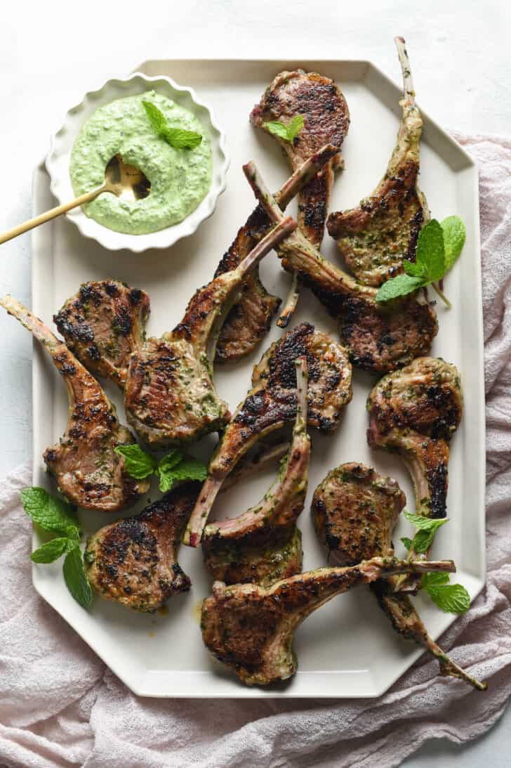 Lamb lollipops on a white geometric platter, garnished with fresh mint, with a small bowl of green dipping sauce.