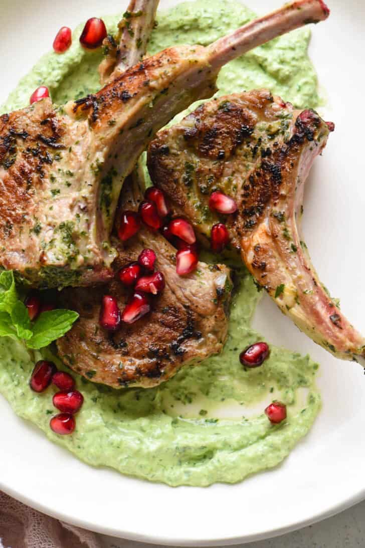 Meaty chops garnished with pomegranate seeds on top of a creamy green sauce, in a white shallow bowl.
