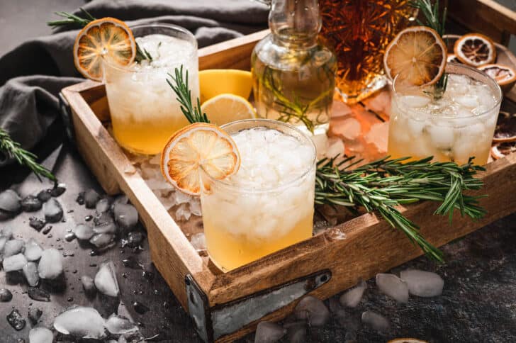 Bourbon lemon cocktails garnished with rosemary and dried citrus on a wooden tray with ice cubes all around the scene.