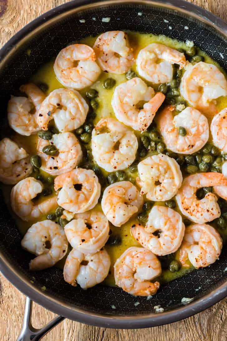 A dark patterned skillet filled with shrimp and capers cooking in butter.