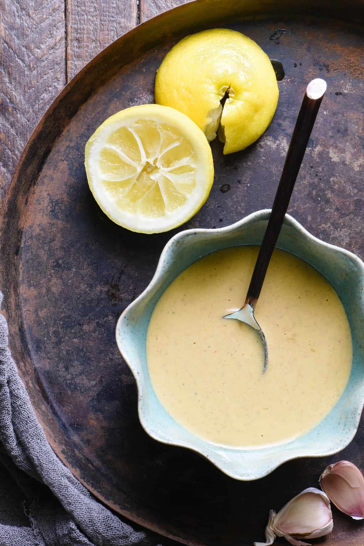 Overhead shot of bowl of tahini dressing with spoon. Lemon halves and garlic cloves on tray with dressing.